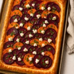 Een goudbruin gebakken plaattaart met dakpansgewijs gerangschikte plakjes zoete aardappel en rode biet, afgetopt met verkruimelde feta en verse peterselie. De plaattaart ligt op een bakplaat met bakpapier en een schaaltje verse peterselie ernaast.
